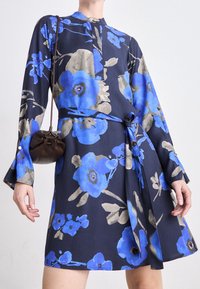 Blommig klänning i marinblått tyg, med stora blå blommor och grå detaljer. Knäppt halsringning och midjeband. En handhållen brun väska ingår.