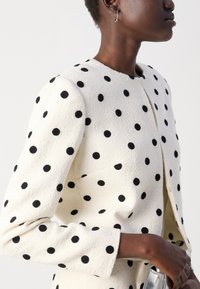 White polka dot jacket with textured fabric, round neckline, structured shoulders, and long sleeves. Features a front closure.