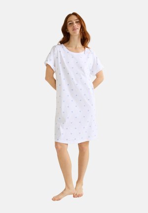 Jeune femme debout pieds nus, portant une robe blanche arrivant aux genoux, à manches courtes et ornée de petits motifs de cœurs bleu clair.