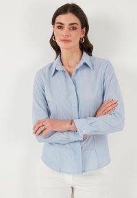 Camicia a righe blu e bianche con bottoni, maniche lunghe, colletto tradizionale e orlo curvo. Il tessuto appare liscio con una texture leggera.