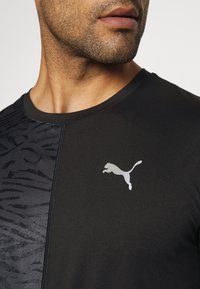 Camisa atlética negra con un patrón texturizado negro en los laterales, con un logo plateado de Puma en el pecho. Material suave y diseño ajustado.