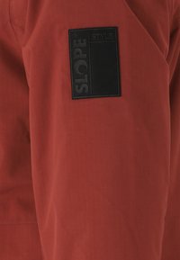 Der rote Oberstoff hat eine strukturierte Oberfläche; ein schwarzes quadratisches Patch zeigt "SLOPE" und "STYLE" in weißer Schrift.
