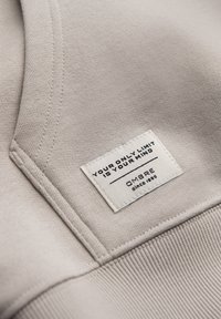 Étiquette en tissu beige sur un vêtement indiquant : « VOTRE SEULE LIMITE EST VOTRE ESPRIT » avec le nom de la marque OMBRE et « depuis 1985 » en dessous.