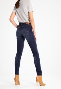 Vaqueros ajustados de color azul oscuro con una cintura alta, que cuentan con bolsillos traseros y una etiqueta de cuero marrón en la parte posterior. Combinados con unos zapatos de tacón altos de color beige.