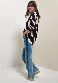 Femme aux longs cheveux bruns portant un pull à motifs noir et blanc, un jean bleu et des baskets beiges, debout de profil les mains dans les poches.