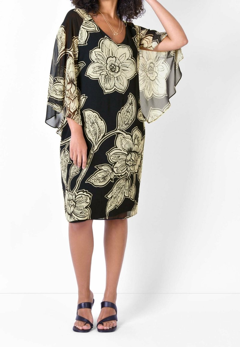 Vestido negro con estampado floral en crema; presenta mangas anchas y fluidas y un diseño de cuello en V. Llevado con sandalias negras.