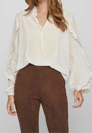 Femme portant un chemisier blanc transparent à manches longues avec des volants et des pois texturés, associé à un pantalon marron taille haute.