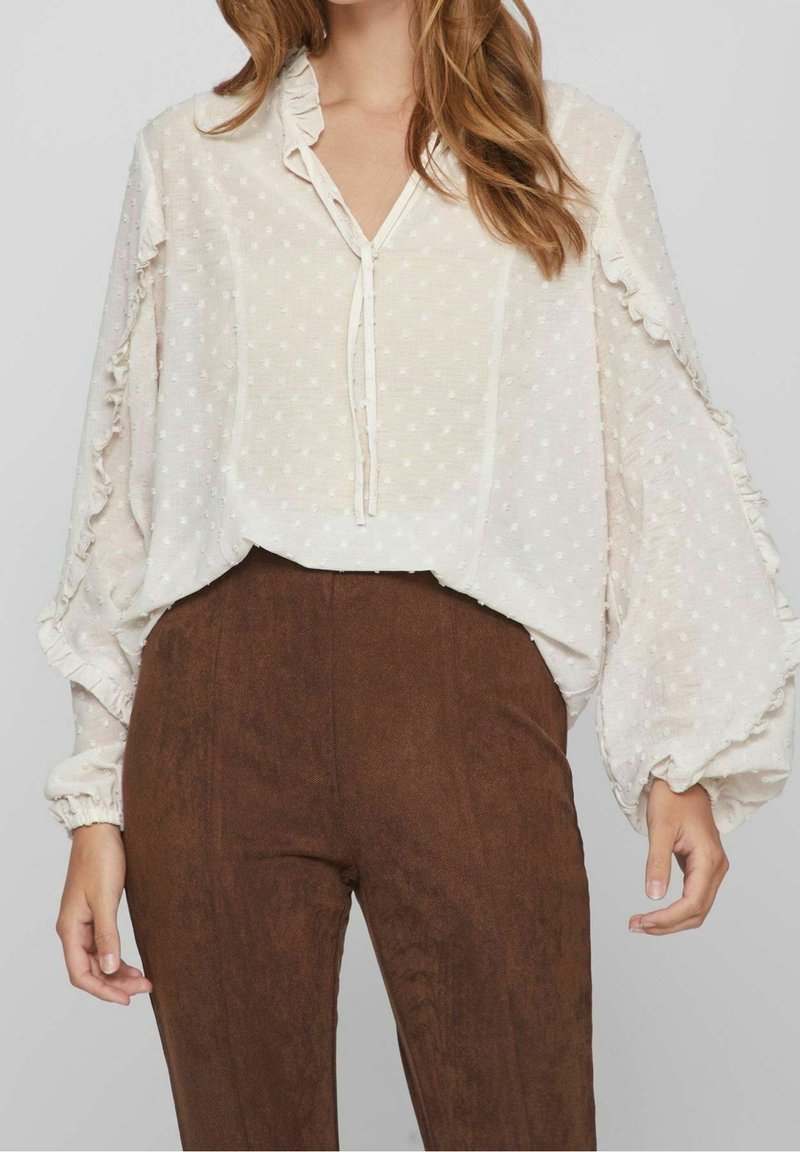 Femme portant un chemisier blanc transparent à manches longues avec des volants et des pois texturés, associé à un pantalon marron taille haute.