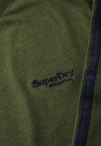 Oliwkowa bawełniana bluza z dekoltem w serek, z ciemnoniebieskimi paskami po bokach i haftowanym logo "SuperDry Athletics".