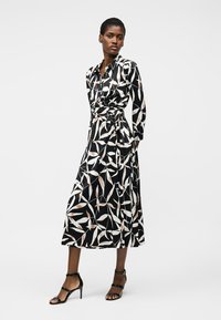 Γυναίκα που φοράει μαύρο midi φόρεμα με λευκά και μπεζ φυλλώδη σχέδια, μακριά μανίκια και μαύρα πέδιλα με τακούνι και ανοιχτά δάχτυλα, στέκεται με τα χέρια πίσω από την πλάτη.