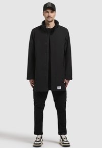 Abrigo negro largo con botonadura frontal, cuello alto y bolsillos laterales, combinado con pantalones cargo y zapatillas negras y blancas. Gorro con logo.