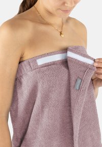 Lila Handtuch aus Frottee mit strukturierter Oberfläche, ausgestattet mit einem weißen Klettverschluss. Ein kleines Stoffetikett ist in der Nähe des Randes sichtbar.