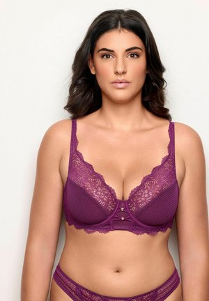 Kvinde med mellembrun hud og mørkt hår iført matchende lilla blonde bh og trusser, vendt fremad mod en ensfarvet baggrund.