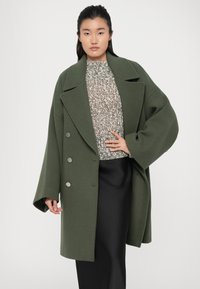Green oversized coat with a shawl collar, double-breasted metallic buttons, and wide sleeves, worn over a patterned knitted sweater and skirt.