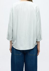 Personne portant une chemise gris clair à manches longues avec des rayures verticales subtiles et un pantalon en denim bleu, vue de dos.