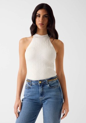 Vrouw met lang donker haar, gekleed in een wit, mouwloos, gestructureerd haltertopje en een blauwe spijkerbroek met een gouden kettingriem, staand tegen een witte achtergrond.