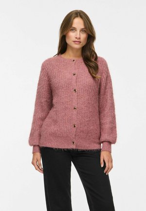 Femme portant un cardigan rose duveteux boutonné et un pantalon noir, debout devant un fond blanc uni.