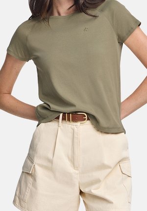 Kobieta ubrana w oliwkowo-zielony T-shirt z krótkim rękawem, włożony do kremowych szortów cargo z wysokim stanem i brązowym paskiem, ręce spoczywają na biodrach.