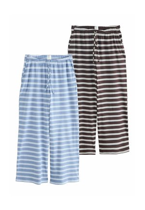Frau steht drinnen, trägt ein schwarzes ärmelloses Top und lockere schwarz-weiße Hose mit horizontalen Streifen, die Hände in den Taschen.