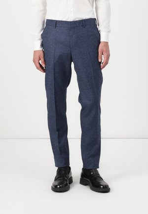 Homme portant un pantalon de costume bleu marine sur mesure, une chemise blanche à manches longues et des chaussures en cuir noir sur un fond uni.