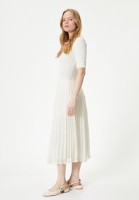 Top bianco a maglia con maniche corte, abbinato a una gonna plissettata color crema con motivo a pois. Il modello indossa scarpe con tacco a blocco beige.