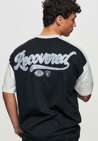 Svart och vit oversized t-shirt med "Recovered" i silver script på baksidan, med sportlogotypdetaljer och korta ärmar.