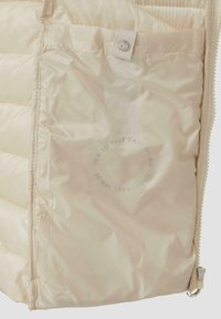 Helle beige Steppjacke mit glatter, glänzender Oberfläche; verfügt über einen Reißverschluss und eine Fronttasche mit einem Pflegeetikett innen.