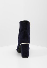 Botines de terciopelo azul marino con tacón bloque, que cuentan con una cremallera lateral y un acento blanco alrededor de la base del tacón.