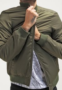 Chaqueta bomber verde oliva con cierre de cremallera, puños y cuello acanalados; llevada sobre una camisa con estampado, que presenta una textura suave y acolchada.