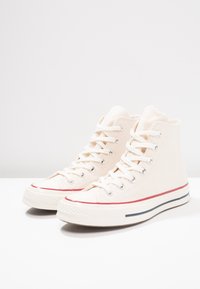 High-top canvas sneakers in off-white with red and blue striped accents, metal eyelets, and flat white laces. Textured rubber sole.