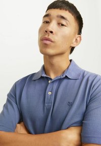 Mladik s kratkom tamnom kosom, odjeven u plavu polo majicu, s prekriženim rukama, samouvjereno gleda u kameru pred jednostavnom pozadinom.