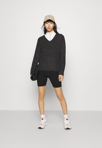Dunkelgrauer V-Ausschnittpullover, schwarze Fahrradhosen, weißes Hemd mit Kragen, beiger Hut, schwarze Handtasche und weiße Sneaker mit roten Akzenten.