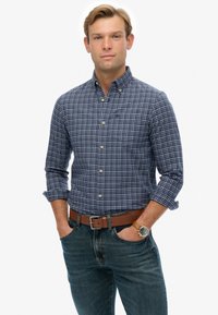 Joven con las mangas arremangadas que lleva una camisa de cuadros azul con botones, jeans, un cinturón marrón y un reloj de pulsera con una correa de cuero marrón.