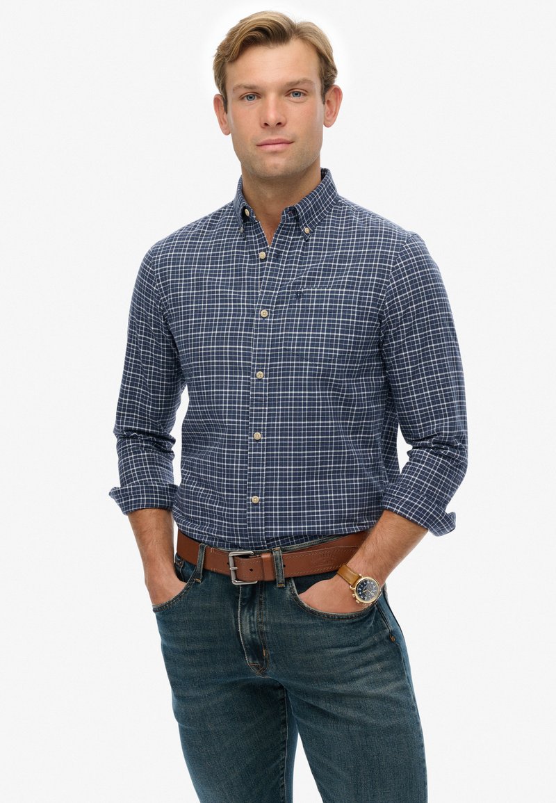 Joven con las mangas arremangadas que lleva una camisa de cuadros azul con botones, jeans, un cinturón marrón y un reloj de pulsera con una correa de cuero marrón.