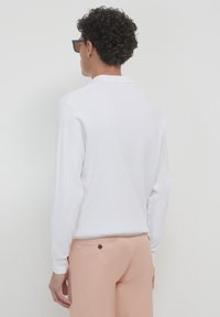 Weißes Langarm-Poloshirt mit Kragen, getragen mit hellpfirsichfarbenen Hose. Das Shirt hat einen strukturierten Stoff und eine taillierte Passform.
