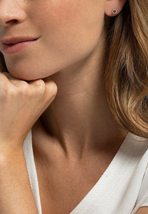 Silberne Steckerohrringe mit einem roten Mittelpunkt, umgeben von klaren Steinen. Das Model hat lange Haare und stützt ihr Kinn auf ihre Hand, während sie ein weißes Oberteil trägt.