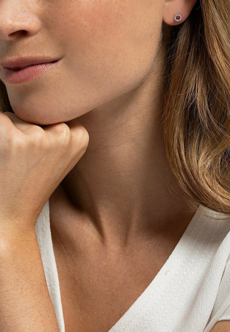 Boucles d'oreilles en argent avec un centre rouge, entouré de pierres claires. Le modèle a les cheveux longs et repose son menton sur sa main, portant un haut blanc.