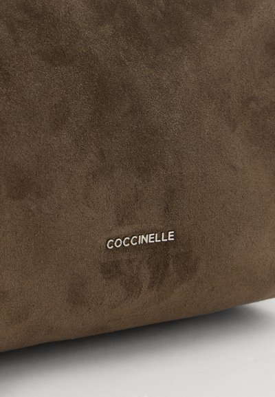 Крупен план на кафява велурена чанта Coccinelle с името на марката, изписано с релефни букви.