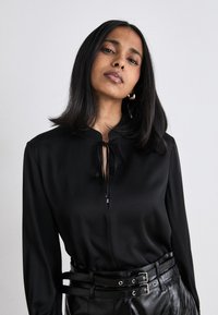 Blusa nera con colletto alto e dettagli con fiocco al collo. Maniche lunghe e tessuto morbido. Abbinata a una cintura in pelle nera e pantaloni.