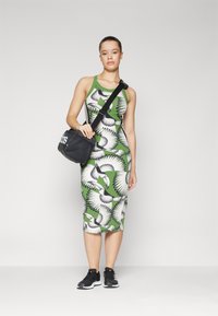 Vestido verde sem mangas com padrão de pássaros em preto e branco, até ao joelho, combinado com sapatilhas pretas e uma bolsa preta com logotipo branco.