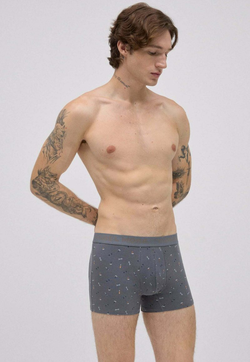 Joven con tatuajes en los brazos y el cuello, vistiendo bóxers grises con un patrón de llaves, de pie contra un fondo claro y liso, mirando hacia abajo.