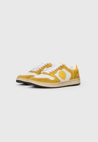 Sneakers amarelas em camurça com detalhes em pele branca, logo redondo e atacadores brancos. Sola em borracha preta. Design atlético clássico.