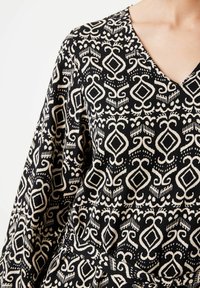 Svart mönstrad klänning med V-ringning, långa ärmar och intrikata beige geometriska mönster på ett lätt material.