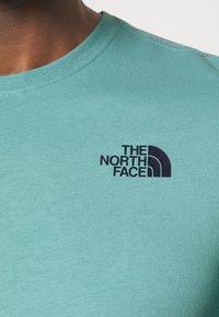 Světle tyrkysové tričko s krátkým rukávem vyrobené z měkké bavlny, s černým logem "The North Face" na levé části hrudi.