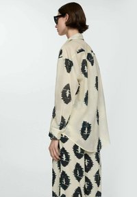 Light beige shirt with black abstract patterns, long sleeves, a relaxed fit, and a curved hem. Paired with matching patterned pants.