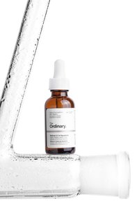 Bouteille compte-gouttes en verre brun étiquetée "The Ordinary" avec un bouchon compte-gouttes blanc, accompagnée d'un morceau de verre clair texturé à côté, les deux recouverts de condensation.