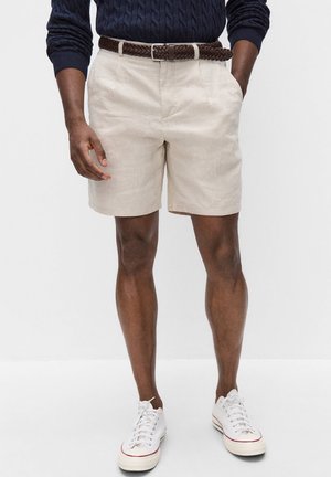 Mand iført beige shorts med et brunt flettet bælte, navy blå kabelstrikket sweater og hvide snørebåndssneakers står mod en hvid baggrund.