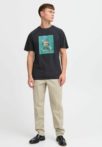 Sort bomulds T-shirt med et farverigt grafik af en dessert på en teal baggrund. Sammen med beige bukser og sorte loafers.