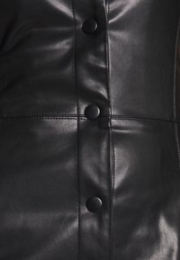 Schwarzes Lederkleidungsstück mit glatter Textur, das sichtbare Nähte und sechs große schwarze Knöpfe entlang der Vorderseite zur Befestigung aufweist.