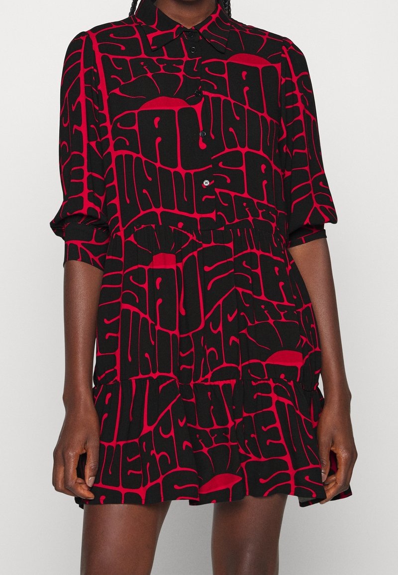 Femme portant une robe noire et rouge à manches longues avec un motif typographique abstrait, audacieux et rouge sur le tissu.